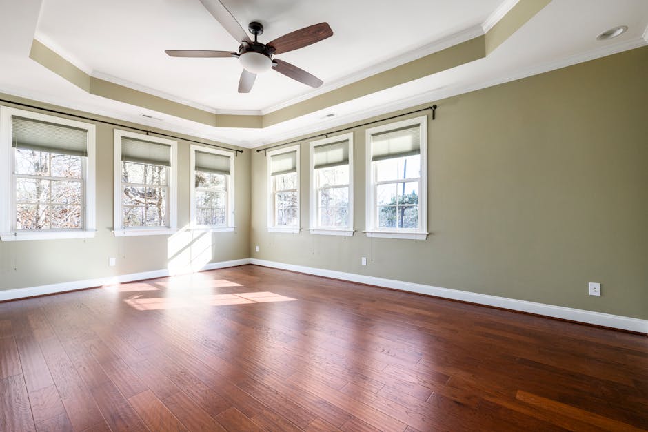 A bright and spacious empty room with large windows, hardwood floor, and ceiling fan.
